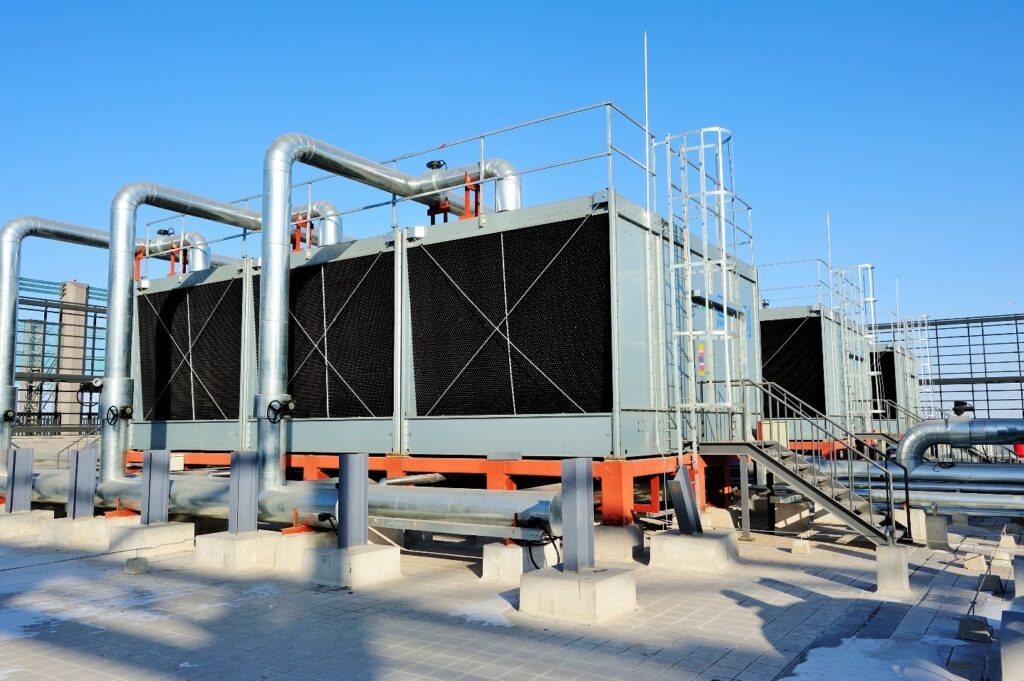 En una azotea, bajo un cielo azul y despejado, hay instaladas grandes unidades de refrigeración industrial con tuberías metálicas y barandillas. Situados sobre plataformas a las que se accede por escaleras, estos sistemas desempeñan un papel fundamental en la reducción de los costes energéticos de las instalaciones.